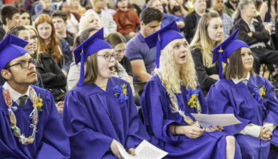 grads wear caps and gowns and sit in front row of an audience during graduation.